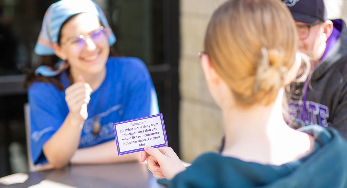 A college student holds a conversation starter card as she facilitates an outdoor community building activity.