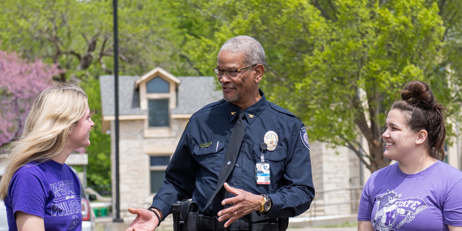 Ronnie Grice, wearing his K-State police uniform, engages with two female students in purple K-State T-shirts outside of the K-State police station.
