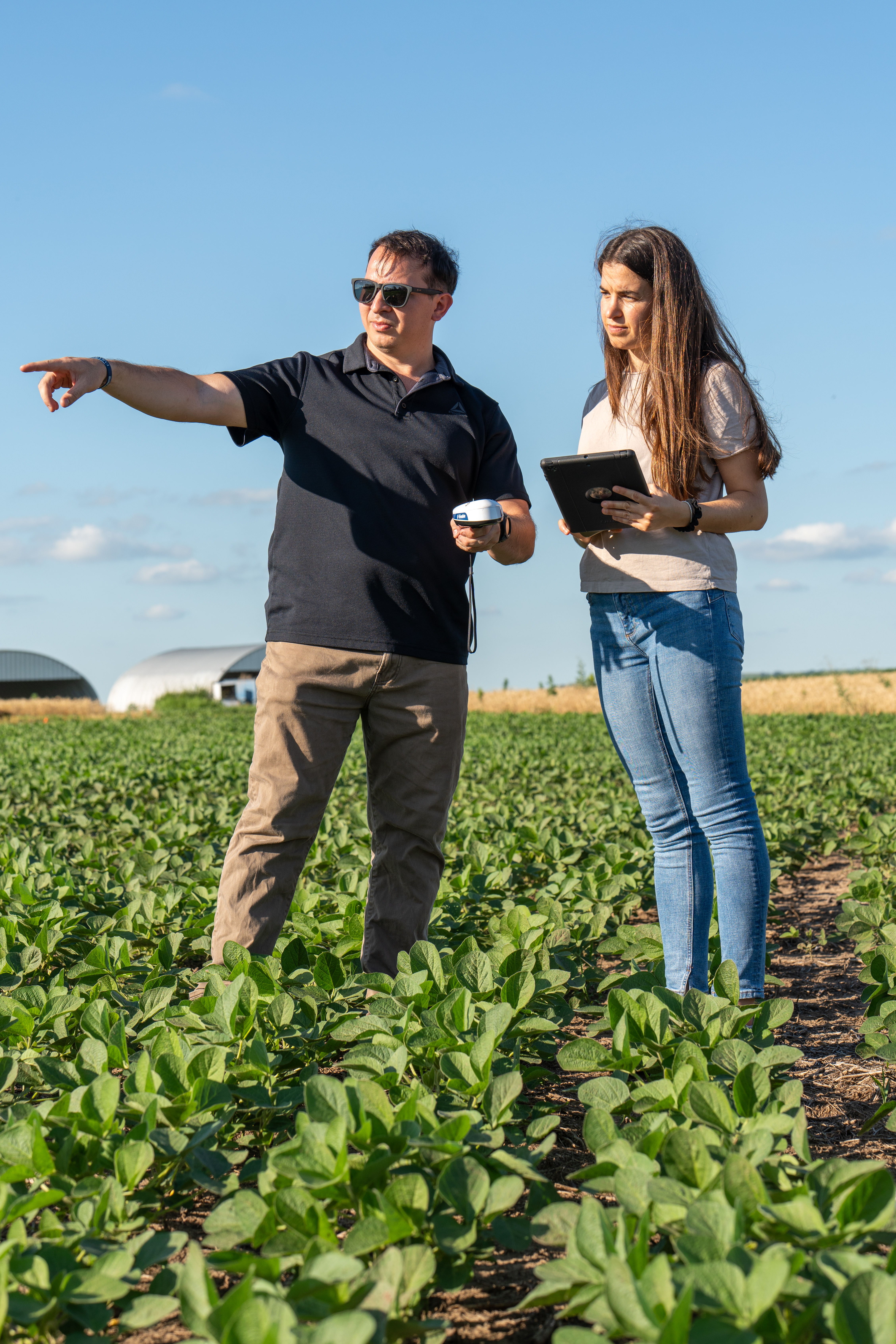 A man wearing a black polo and khaki pants stands with a young woman wearing blue jeans and a white shirt in a soybean field. The man is pointing down the field while holding a white piece of technology, and the woman is holding a tablet.