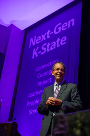 A man in a suit and tie smiles in front of a purple Next-Gen K-State backdrop