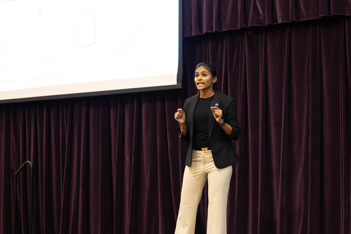 A doctoral candidate in a black blazer and white pants presents a research presentation on a stage on a ballroom.
