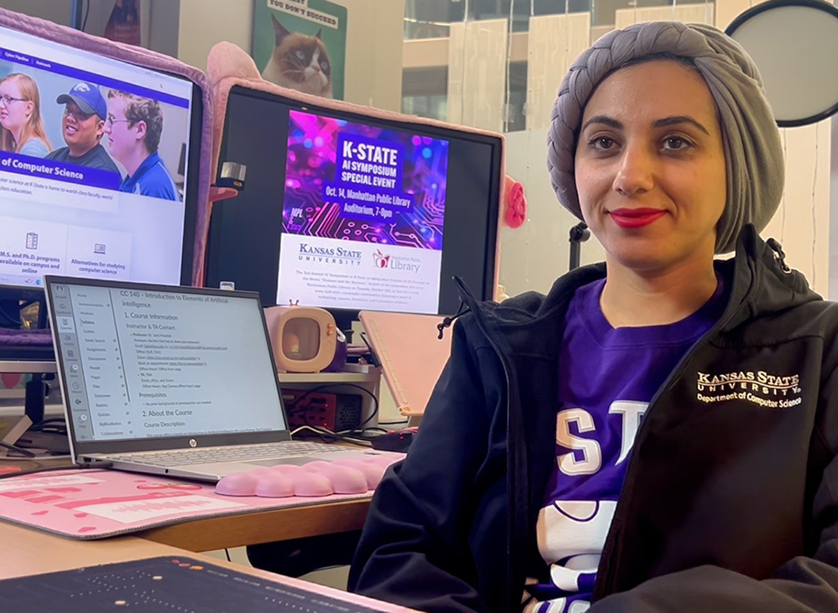 A woman in a black K-State Computer Science sweater and a gray head wrap sits next to three computer screens showcasing different computer science websites and presentations.