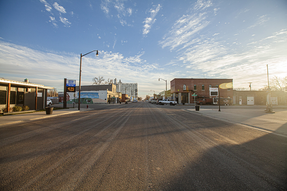 A wide shot of the rural town of Courtland, Kansas, showing the road in the foreground and a blue sky filled with clouds in the background, with a Courtland business block shown in the middle of the photo.