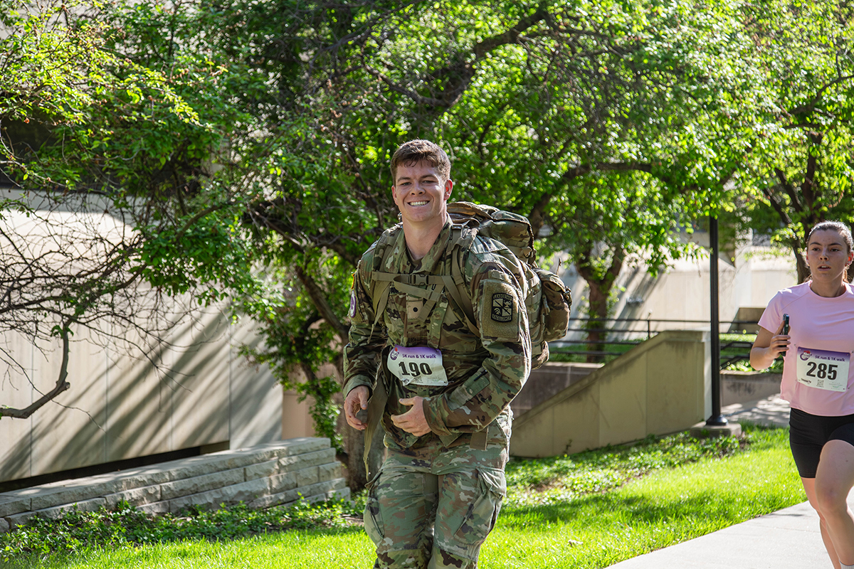A smiling Kansas State University Army ROTC cadet wearing full military gear and a rucksack runs through a tree-lined campus path during the Run for the Roses 5K.