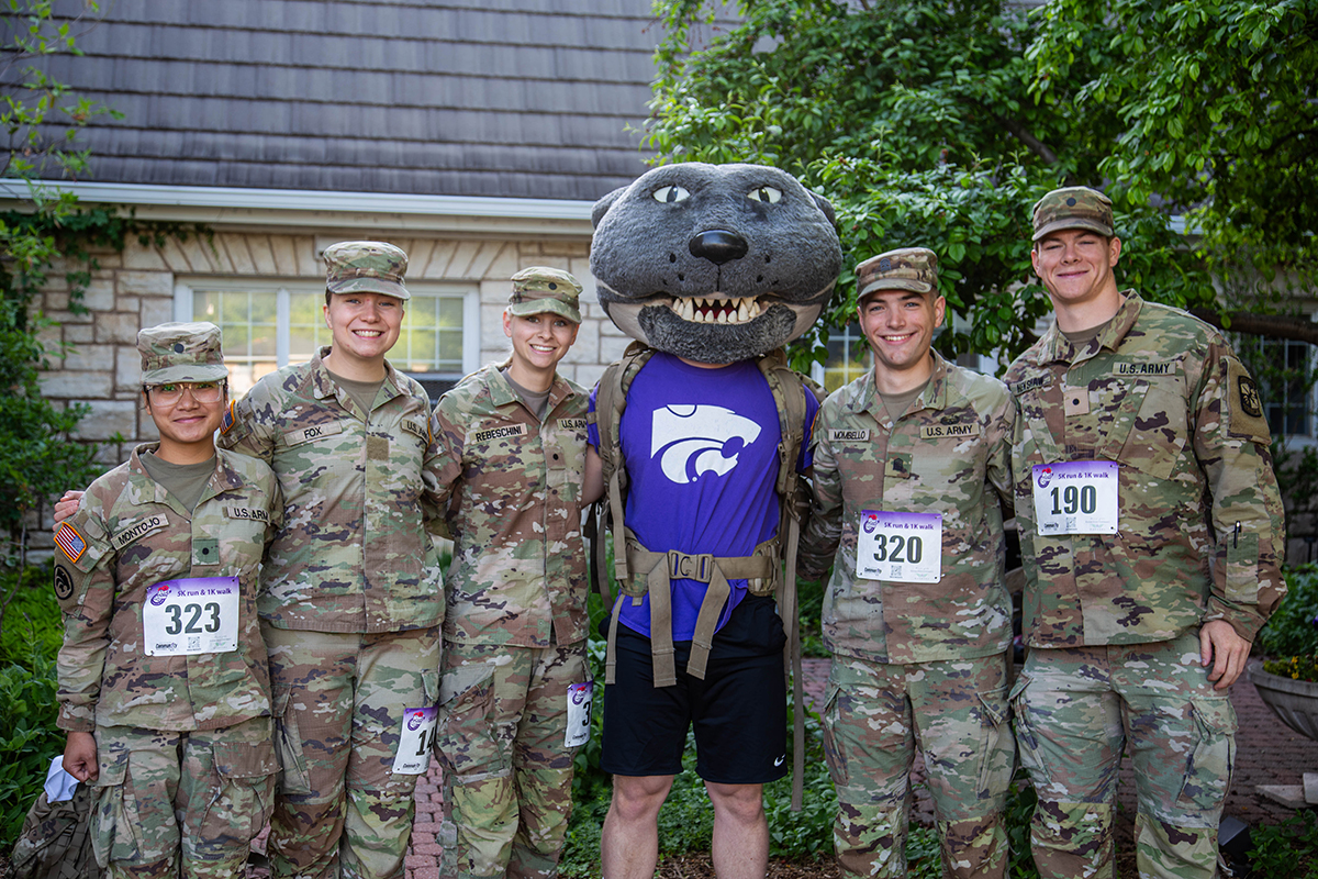 Five Kansas State University Army ROTC cadets in camouflage uniforms and race bibs pose with Willie the Wildcat mascot wearing a purple K-State jersey in front of a stone building with green foliage.