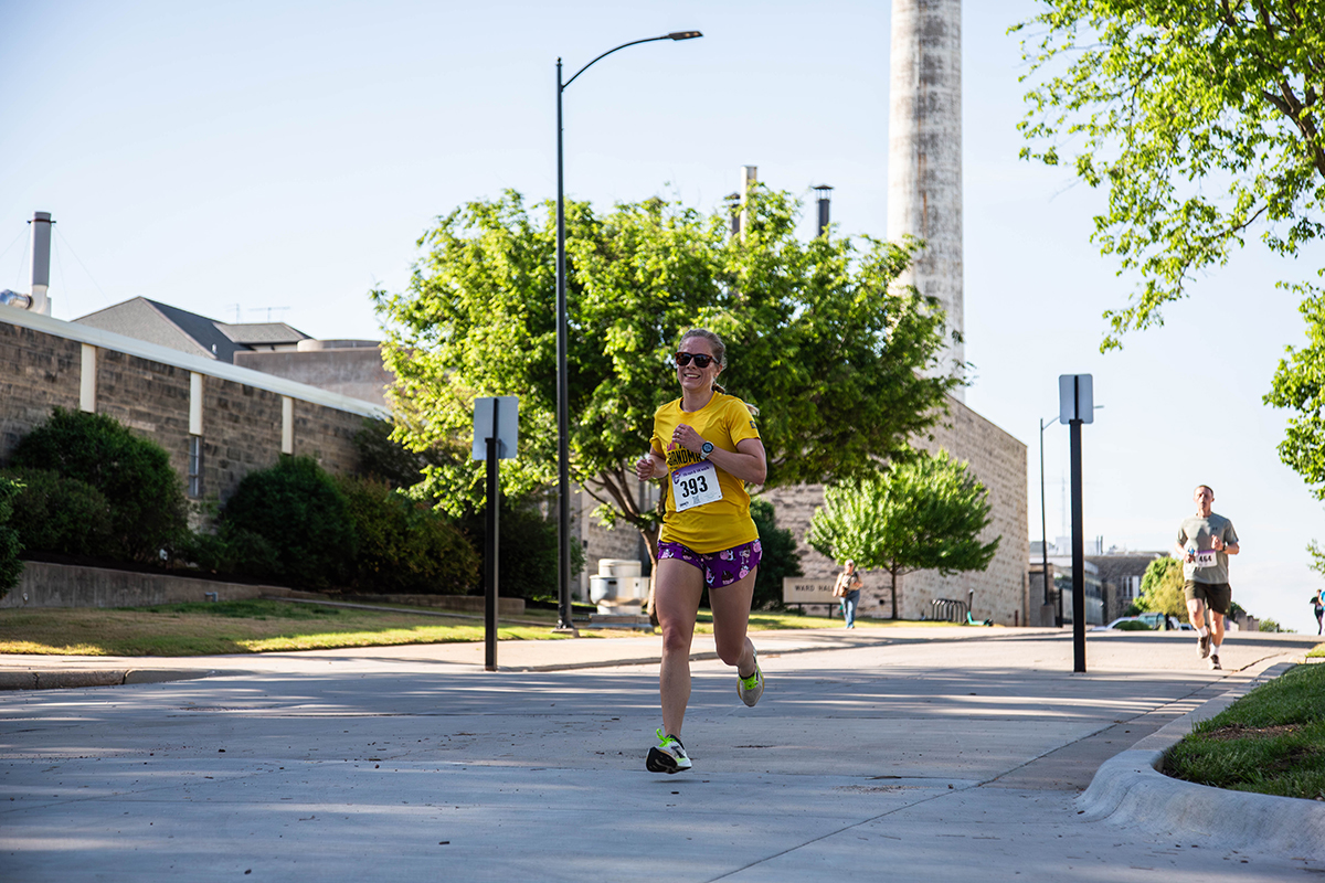 A female runner wearing a yellow shirt, purple shorts, sunglasses and race bib number 393 runs down a city street past industrial buildings and green trees during the Run for the Roses 5K with another runner visible in the background.
