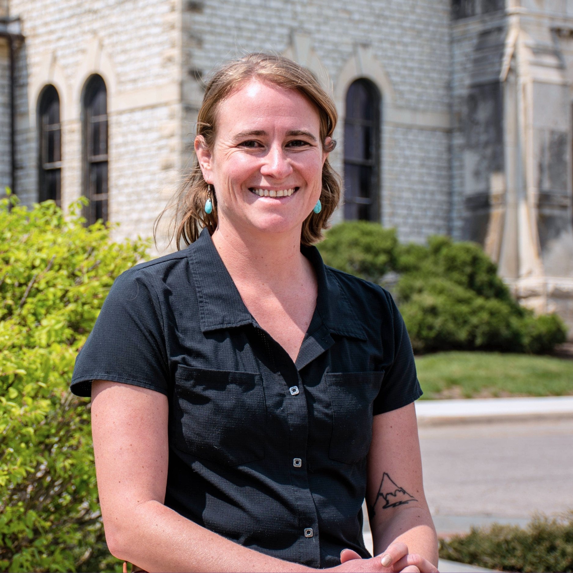 Morgan Greene smiles for a photo in front of Anderson Hall. She wears a black short-sleeve button-up shirt and turquoise earrings. A tattoo of a mountain is visible on her forearm.