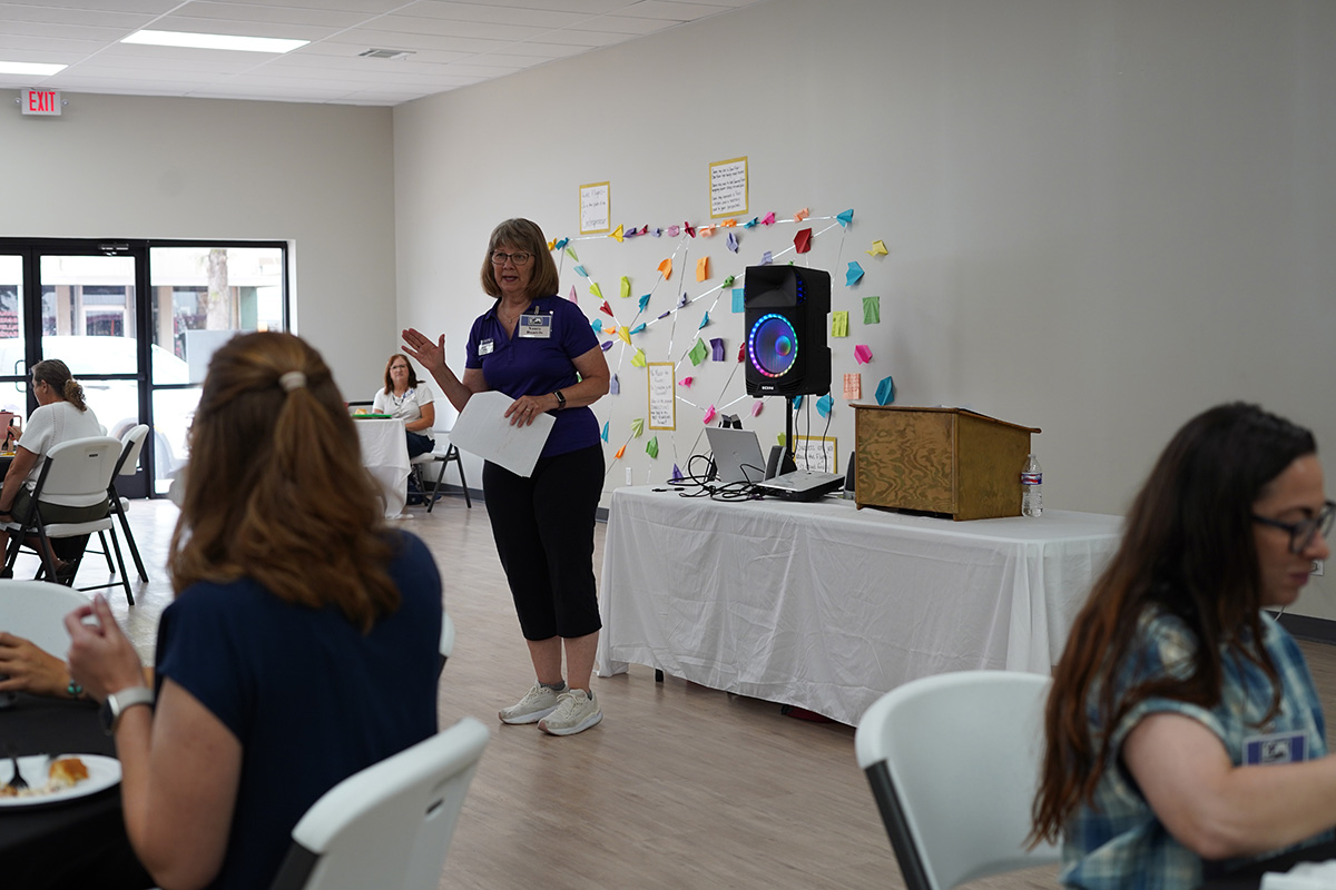 A woman stands in a community gathering room and addresses the audience with presentation boards behind her. 
