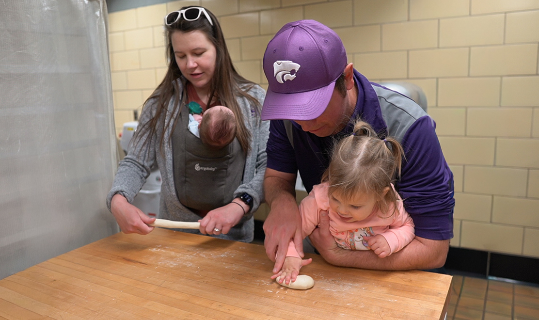 ALT Text: A family makes dough together in a Kansas State campus kitchen, with an adult wearing a purple K-State Wildcats cap helping a young child while another adult with a baby in a carrier rolls dough.