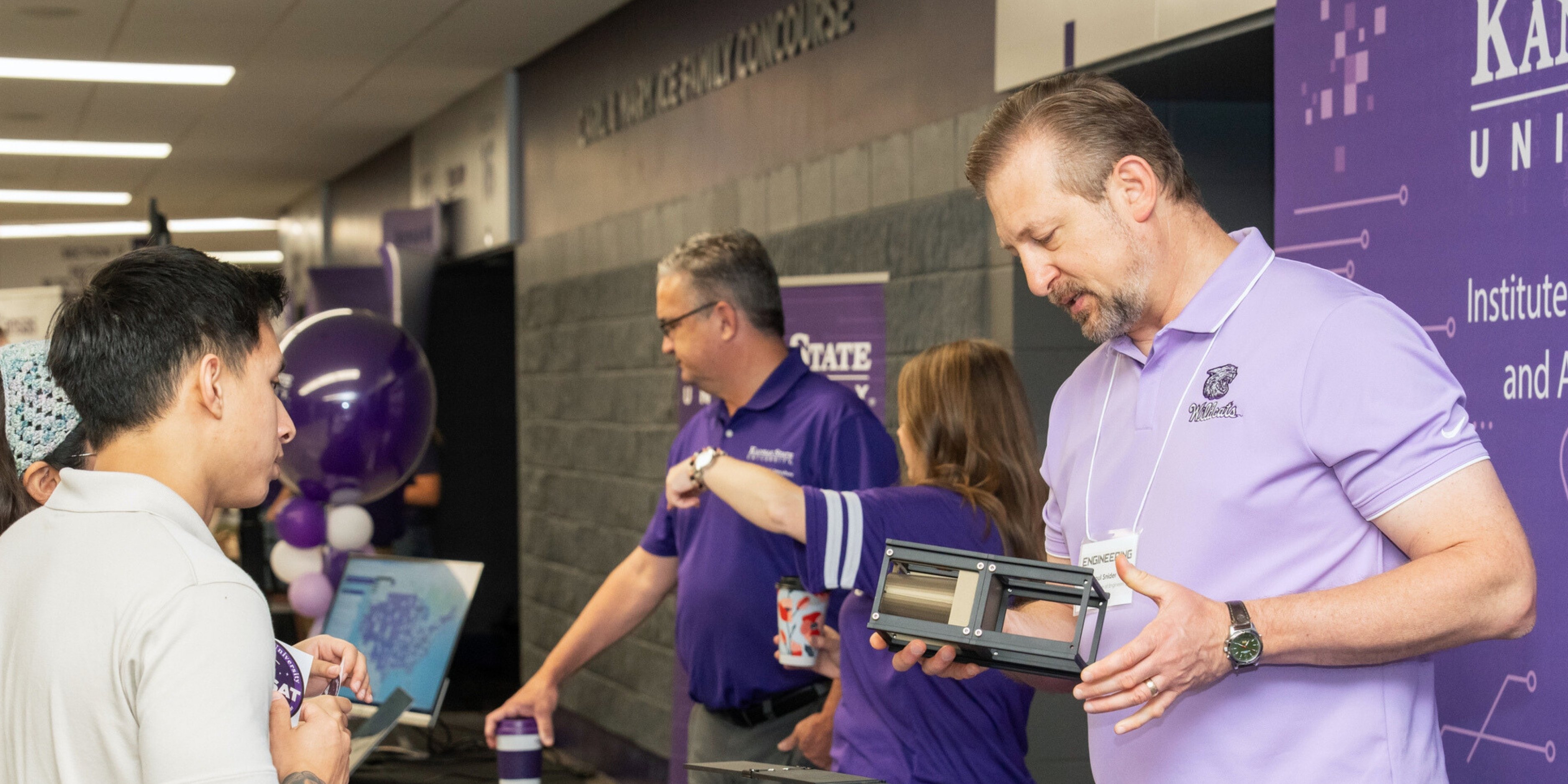 Paul Snider, in a lavender polo, stands at a table in Bramlage Coliseum for the Institute for Digital Agriculture and Advanced Analytics. Behind him is a purple pull-up banner. He speaks with two people: a man in a white polo and a woman wearing a blue crocheted headband.