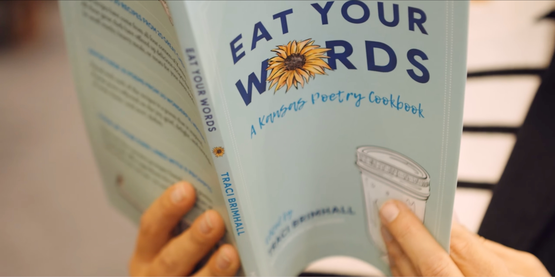 Close up of Brimhall's book. The cover is a light blue, with the title, "Eat Your Words: A Kansas Poetry Cookbook," displayed on the front cover. The "O" in "Words" is replaced with a yellow sunflower.