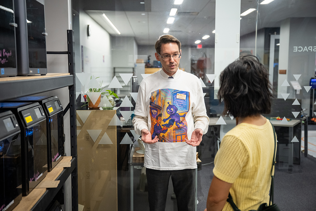A man in a white long-sleeved shirt speaks to a woman in a yellow shirt in front of some 3D printers