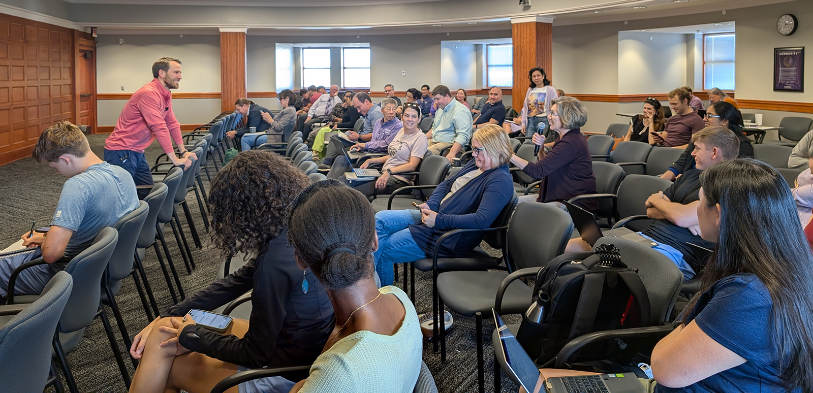 A group of people seated in chairs watch as a woman in the audience asks the presenter a question.