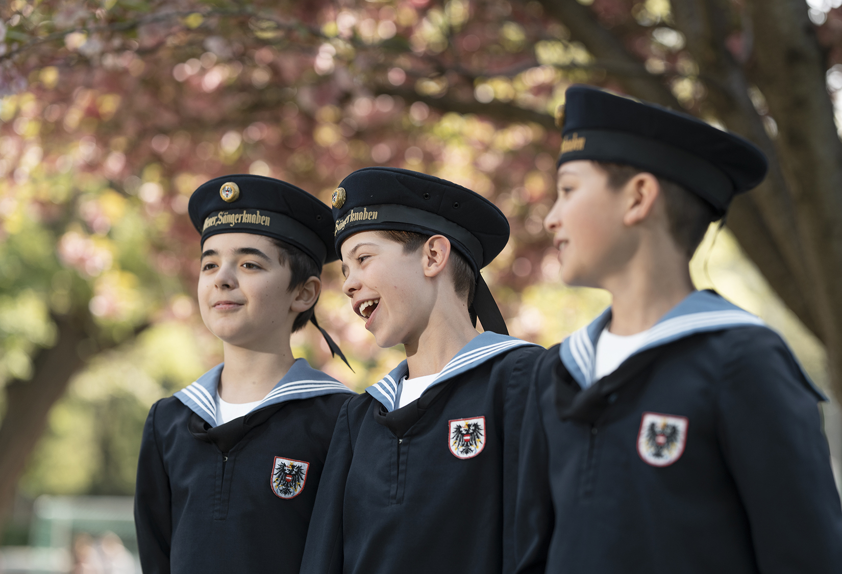 Three boys in navy-colored uniforms with hats smile and sing in front of a tree that is blurry in the background with red-pink leaves.