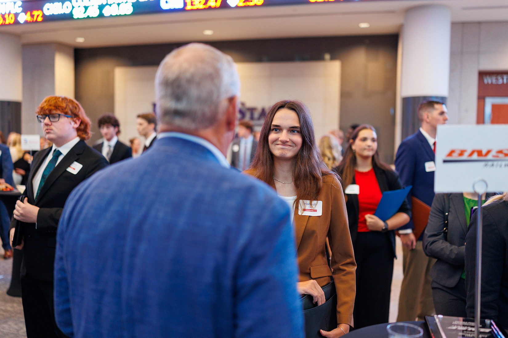 A student in a tan blazer interacts with a corporate partner, a man in a blue suit with his back to the camera.