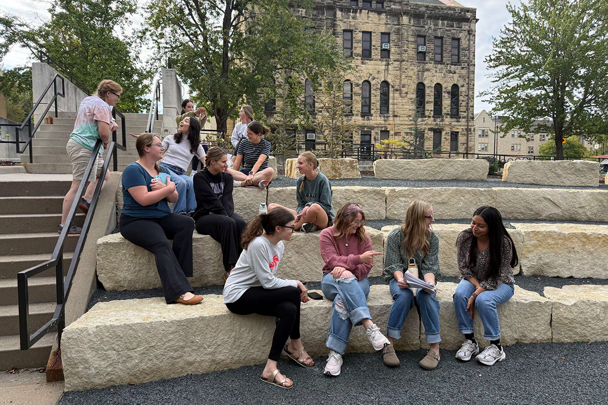 A group of college students sit on limestone blocks as part of an outdoor ampitheater on a college campus.