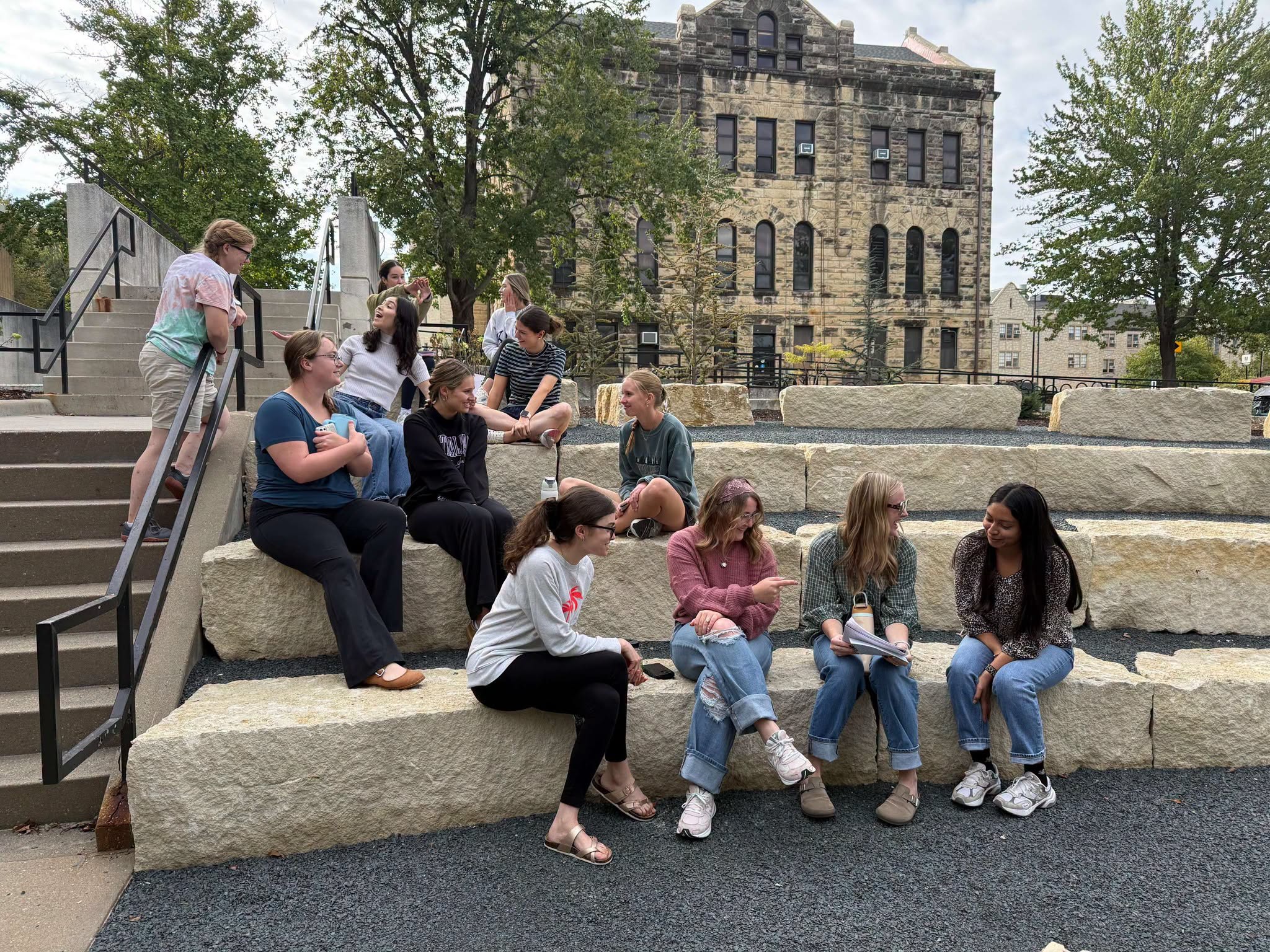 A group of college students sit on limestone blocks as part of an outdoor ampitheater on a college campus.