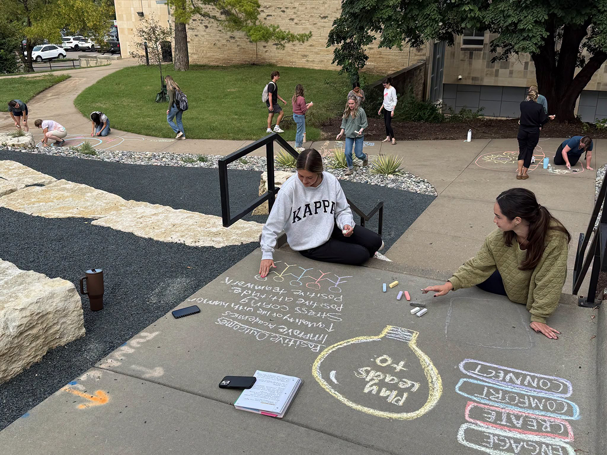 Students write on chalk at an outdoor amphitheater space on a college campus.