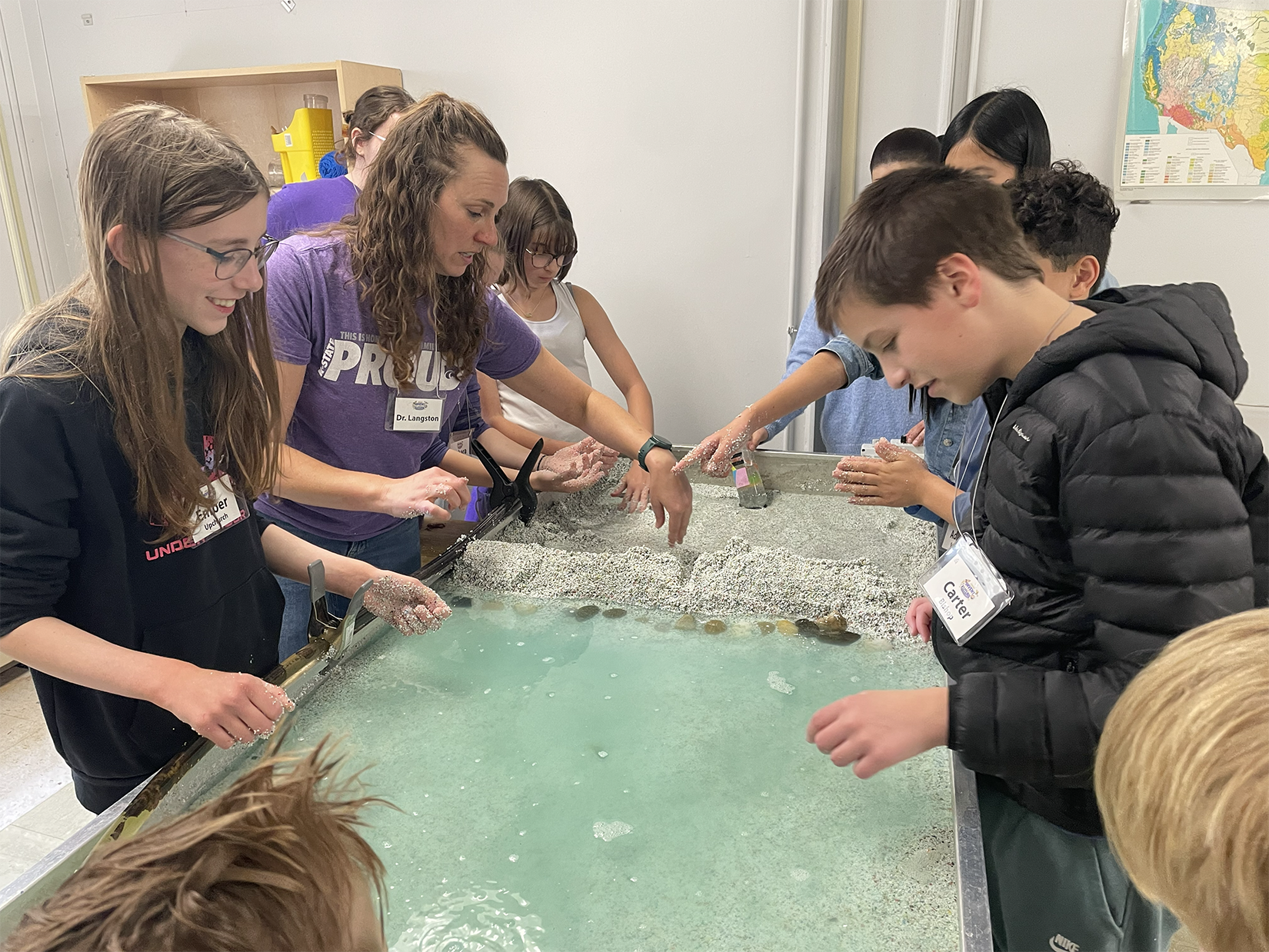 A woman in a purple t-shirt and middle school students stand around a hands-on activity that features water, rocks and sand.