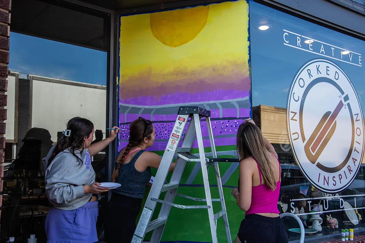 A group of female student painting a stadium onto a window.
