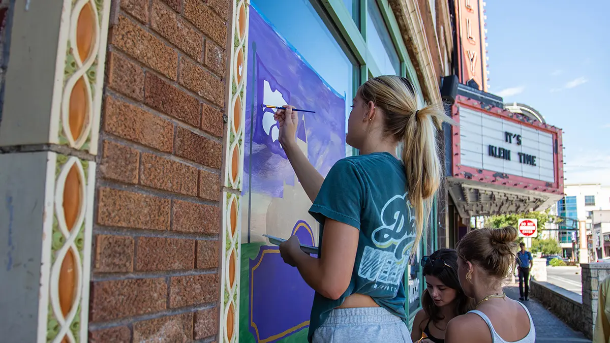A female student painting the Wildcat logo into a window of Rally House.