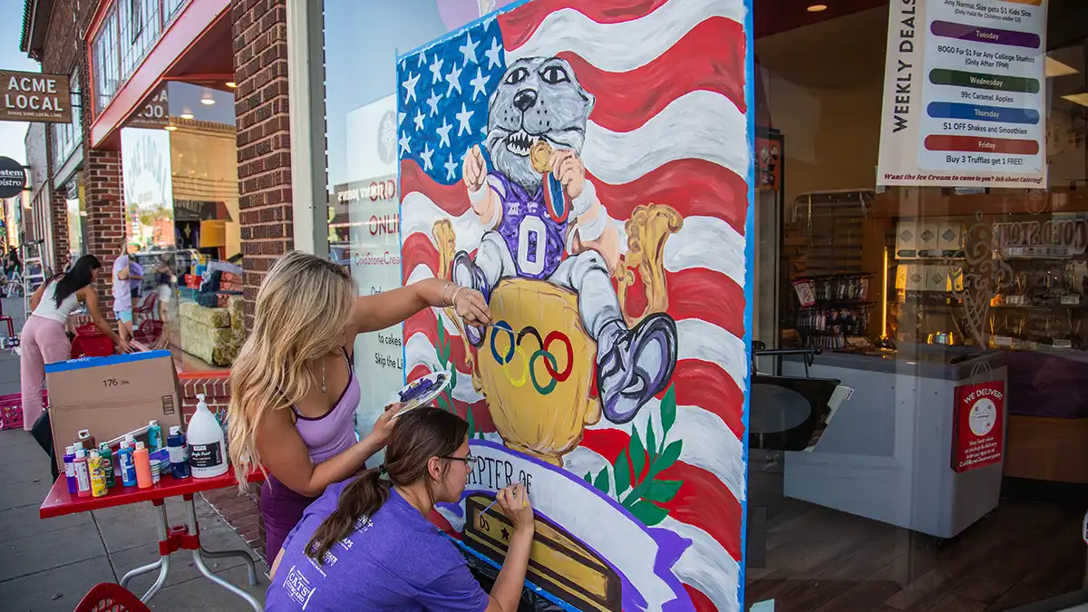 Two female college. students paint a mural of a purple wildcat mascot in front of an American flag on a storefront's window.