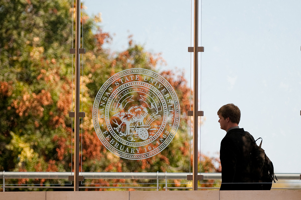 A college student walks past a glass window with the Kansas State University seal embossed on the window. A large tree with fall foliage is in the background. 