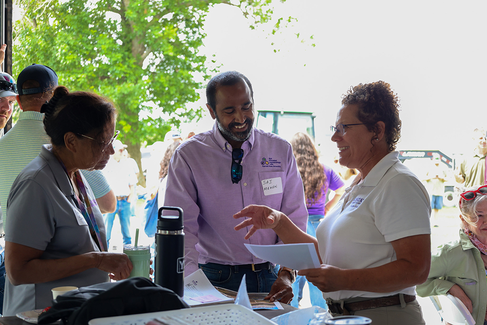 Three people stand around an outdoor high-top table gesturing to papers and smiling