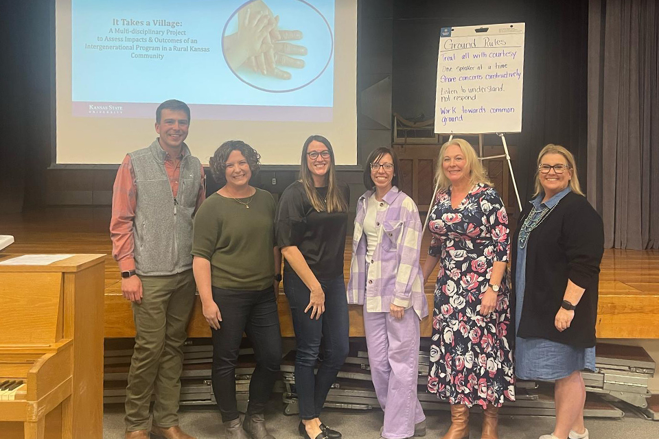 A group of researchers stand in front of a stage. Behind them on a screen, a slide titled "It Takes a Village: A Multidisciplinary Project to Assess Impacts and Outcomes of an Intergenerational Program in a Rural Kansas Community" is presented.