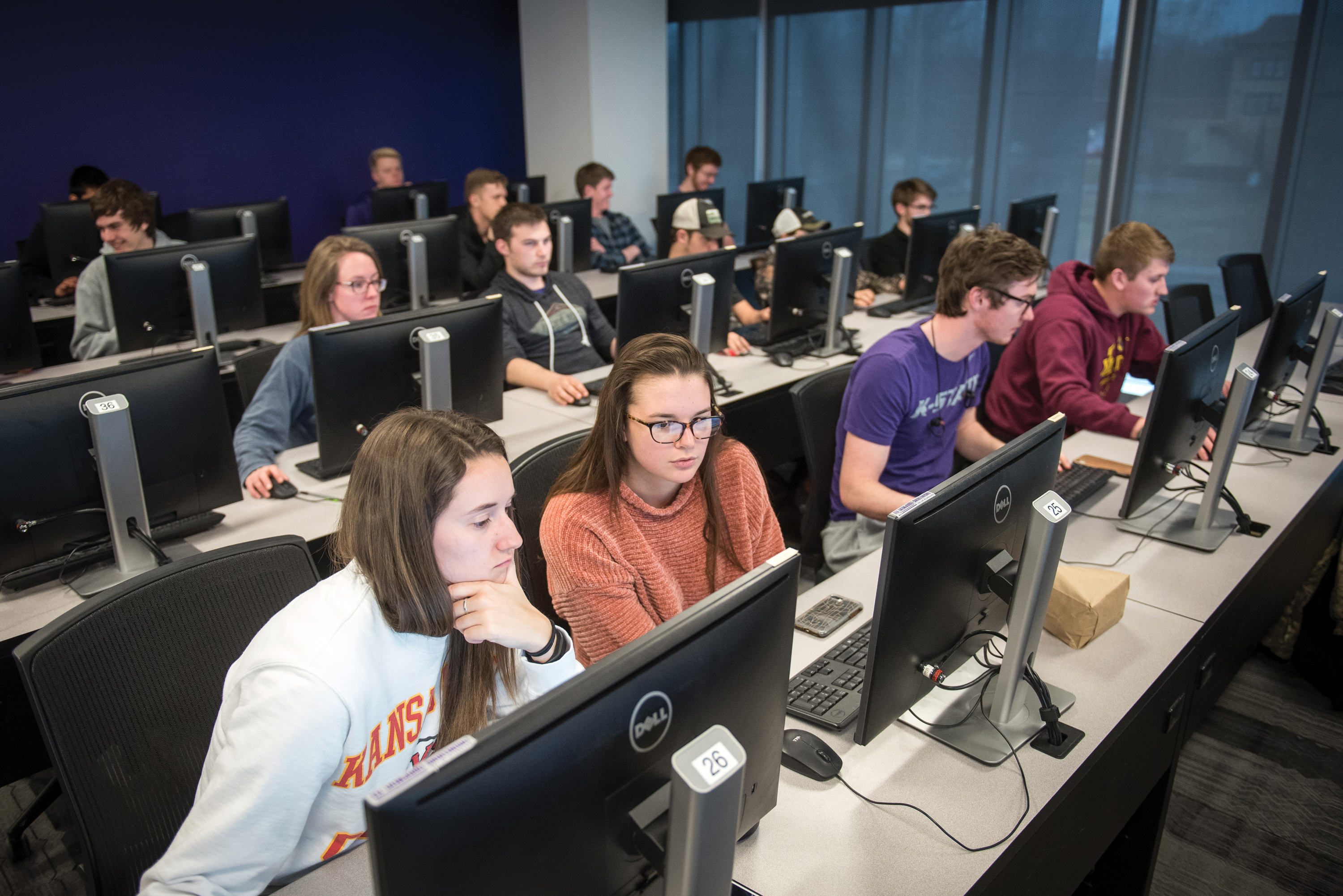 In several staggered rows of desks, students sit at computers and work on computer science skills.