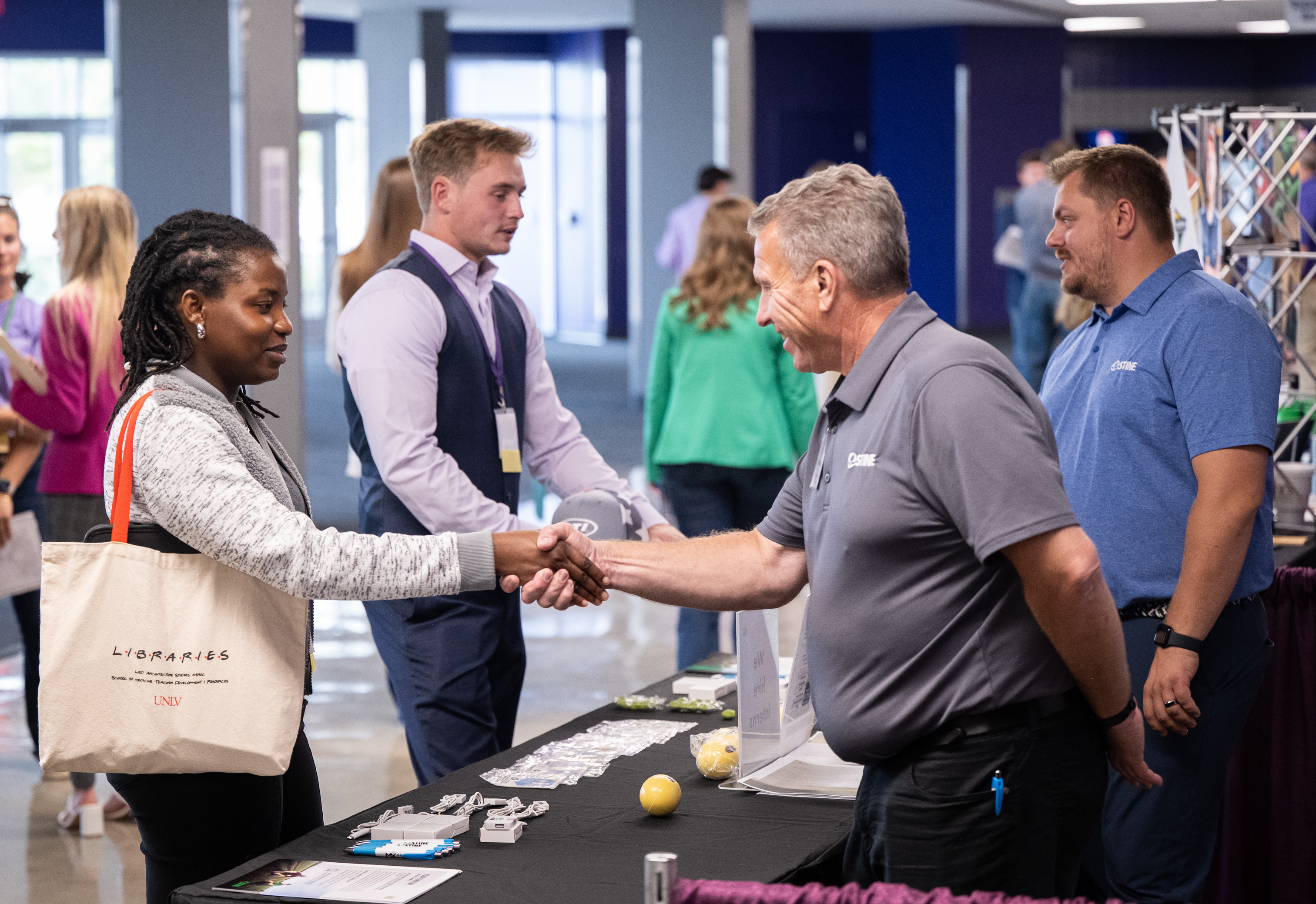 Two K-State students talking with employers at a career fair. Man in gray shirt has his hand out to shake hands with Black female student.