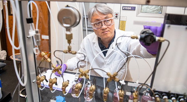 A man in a white labcoat and safety glasses works with tubing.