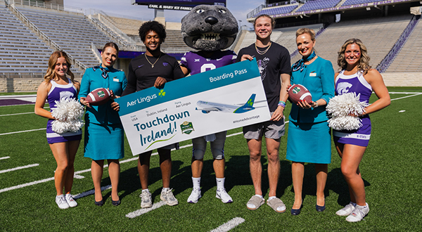 A college mascot of a Wildcat stands on a football field and holds a large, mock boarding pass. Several football players, cheerleaders and flight attendants stand beside him.