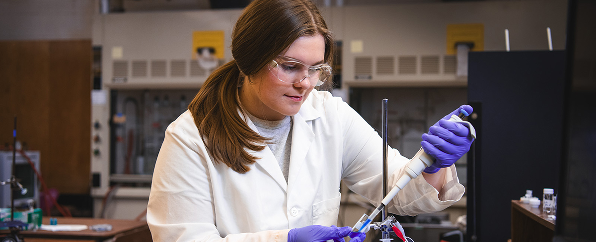 A young woman in a white lab coat and purple gloves works with lab equipment.
