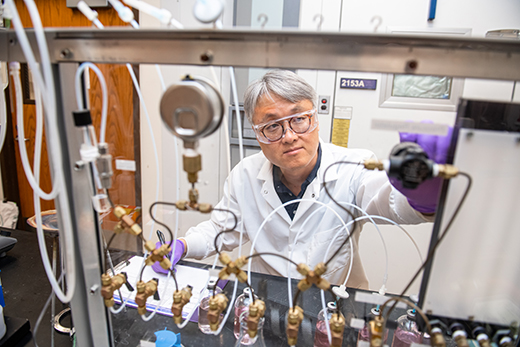 Sample Wearing a white lab coat and safety goggles, Jeongdae Im sits behind and adjusts a row of hoses at a laboratory work bench.