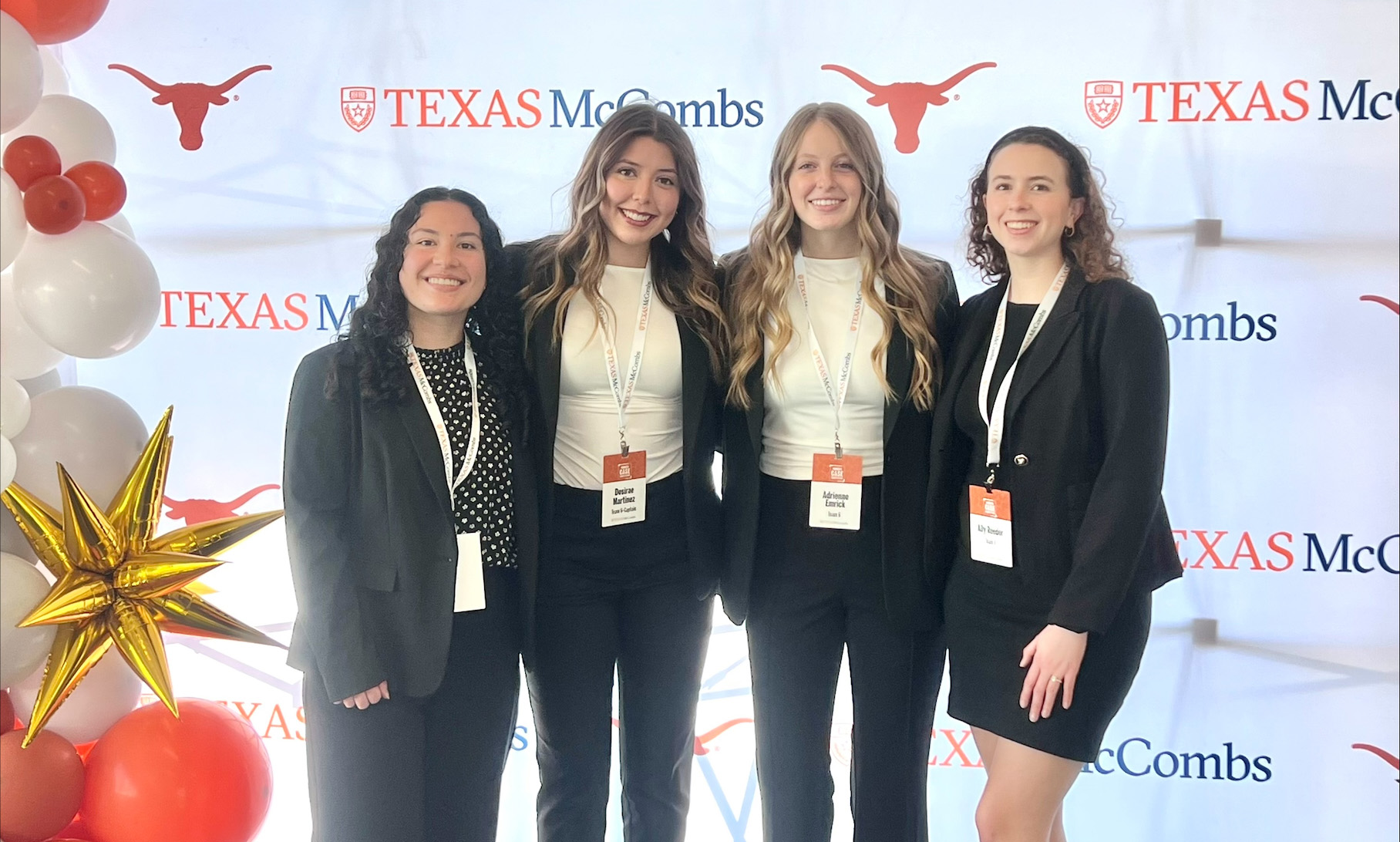 Four young women in professional attire stand in front of a white banner with Texas longhorn symbols and the name of the marketing competition, TexasMcCombs, near an orange and white balloon structure.